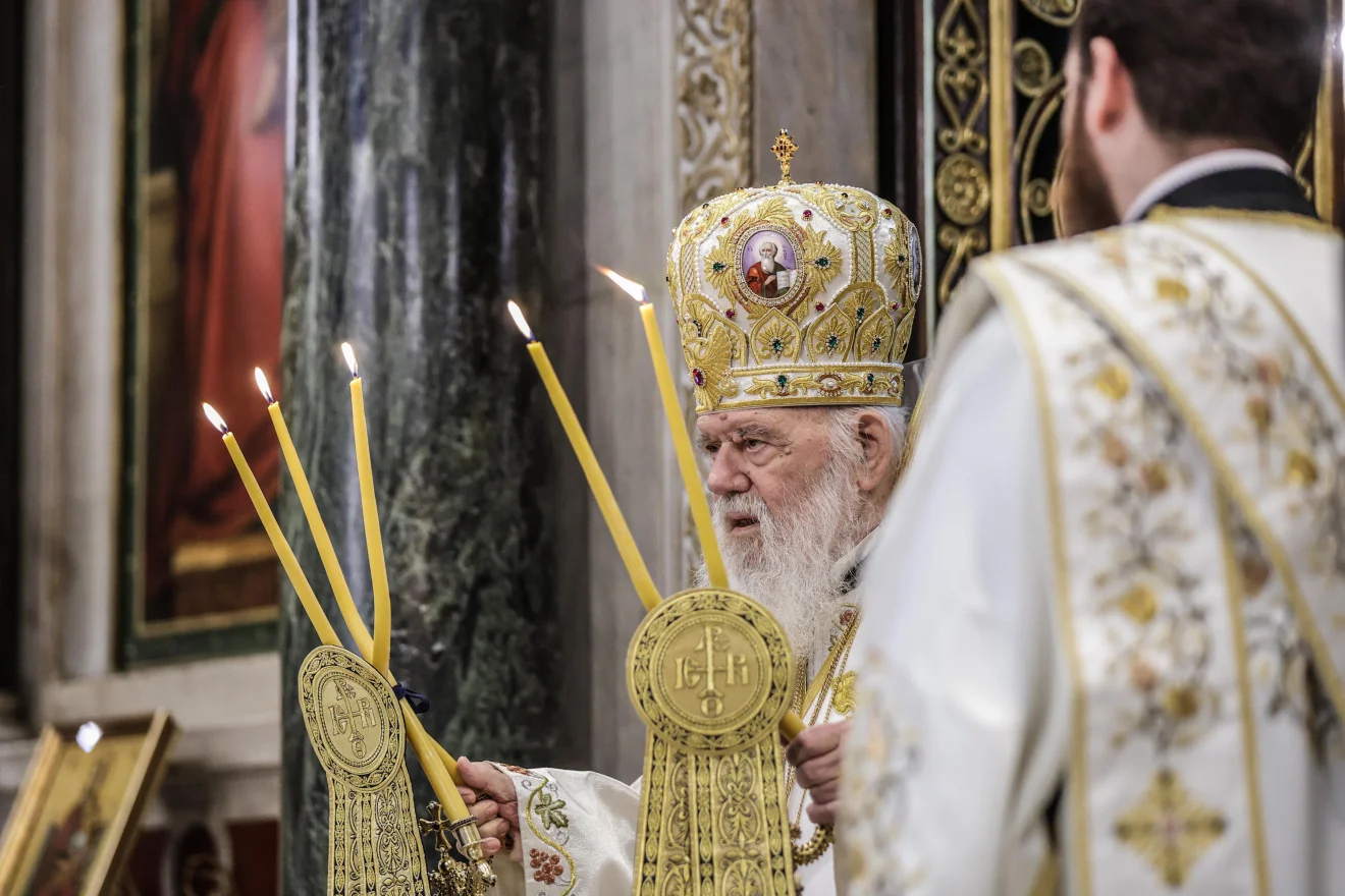 Ο Αρχιεπίσκοπος Ιερώνυμος τελεί θεία λειτουργία, φορώντας επίσημα άμφια και μιλώντας μπροστά στα μικρόφωνα. Η εικόνα αποτυπώνει κατανυκτική στιγμή μέσα στον ναό, με αναμμένες λαμπάδες στο προσκήνιο.  Πηγή: INTIME