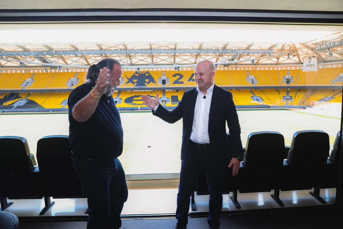 Δύο άνδρες συνομιλούν σε σουίτα της OPAP Arena, με τις κερκίδες της ΑΕΚ να φαίνονται στο φόντο. Πηγή: ΙΝΤΙΜΕ.