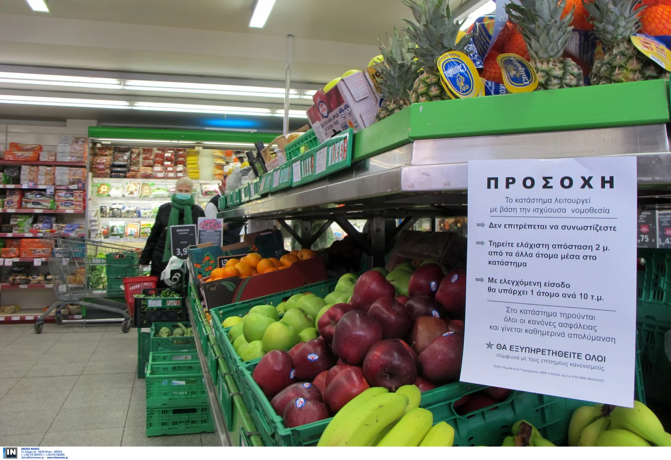 Καταναλώτρια με μάσκα κάνει τις αγορές της σε σούπερ μάρκετ, δίπλα σε πάγκο με φρούτα και ενημερωτική σήμανση για μέτρα προστασίας. Πηγή: ΙΝΤΙΜΕ.