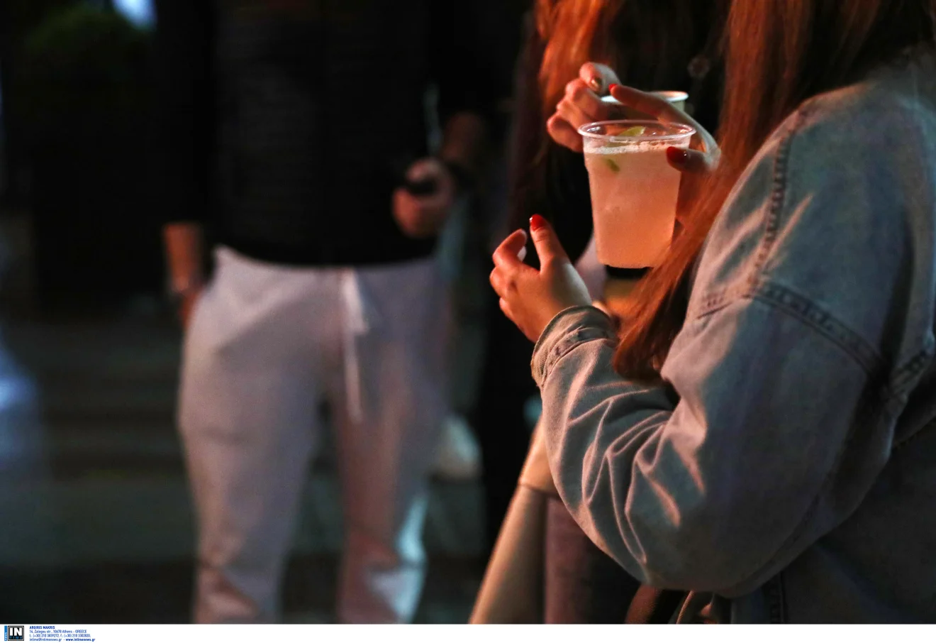 Νεαρή γυναίκα κρατά ποτό σε πλαστικό ποτήρι, σε νυχτερινό στιγμιότυπο διασκέδασης με χαμηλό φωτισμό. Πηγή: ΙΝΤΙΜΕ.