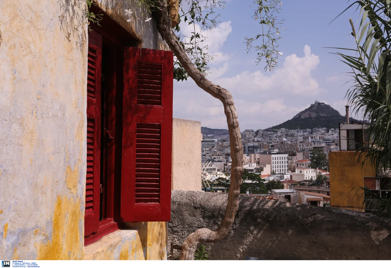 Παραδοσιακό σπίτι με κόκκινα ξύλινα παντζούρια και αναρριχώμενη βλάστηση, με θέα προς την πόλη και τον Λυκαβηττό. Πηγή: INTIME.