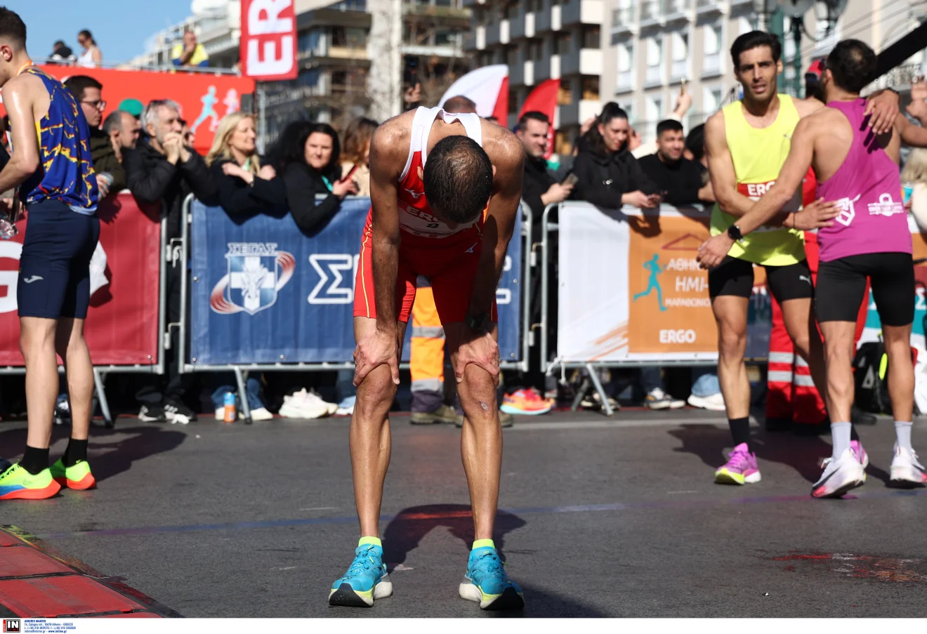 Δρομέας σκύβει εξαντλημένος στον τερματισμό αγώνα δρόμου, ενώ άλλοι αθλητές και θεατές βρίσκονται γύρω του. Πηγή: INTIME.