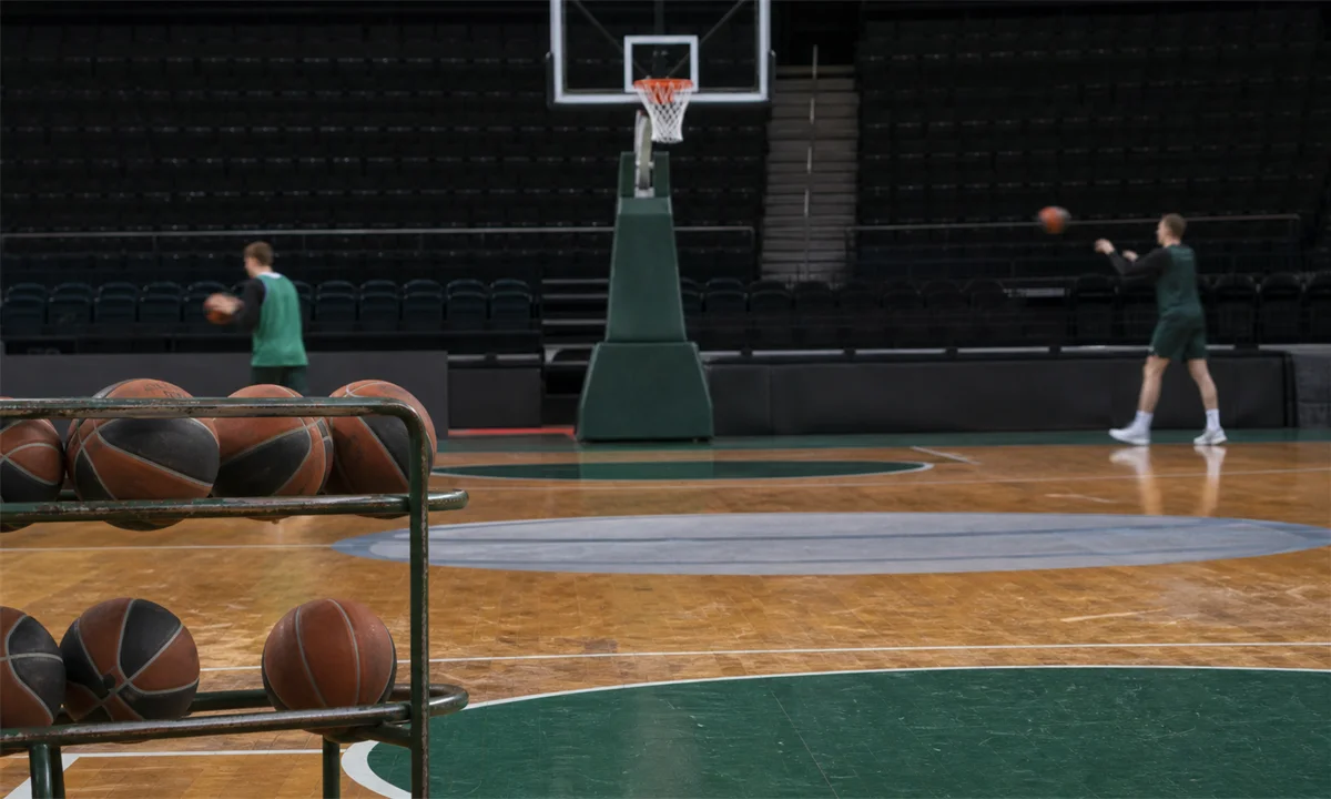 Κλειστό γήπεδο μπάσκετ με ράφι από μπάλες, φθαρμένο παρκέ και δύο μακρινούς παίκτες σε προπόνηση – Πηγή: Sportime.gr